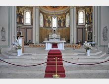 Innenansicht einer Kirche mit einem erhöhten Altarbereich. Der weiße Altar steht auf einem Podest mit einem roten Teppich, umgeben von Kerzen und Blumen; dahinter eine ornamental verzierte Apsis mit goldenen und religiösen Wandmalereien, flankiert von Statuen und Kirchenfenstern.