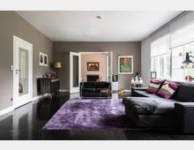 Modernes Wohnzimmer mit dunklem Holzboden, lilafarbenem Teppich und einer grauen Sitzlandschaft. Die Einrichtung umfasst ein Sideboard mit Dekoration, große Fenster mit Jalousien und einen Übergang zu einem weiteren Raum im Hintergrund.