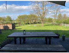 Holztisch und Bank auf einer Terrasse im Vordergrund, mit Blick auf eine weitläufige Wiese, einzelne Bäume und einen Zaun im Hintergrund. Unter blauem Himmel sind ländliche Elemente wie ein Holzschuppen und Gartenaccessoires sichtbar.