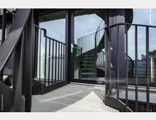 Metallene Außentreppe mit Gittergeländer führt zu einer Glastür, hinter der eine innenliegende Wendeltreppe sichtbar ist. Im Hintergrund sind Gebäude und ein wolkiger Himmel zu erkennen.