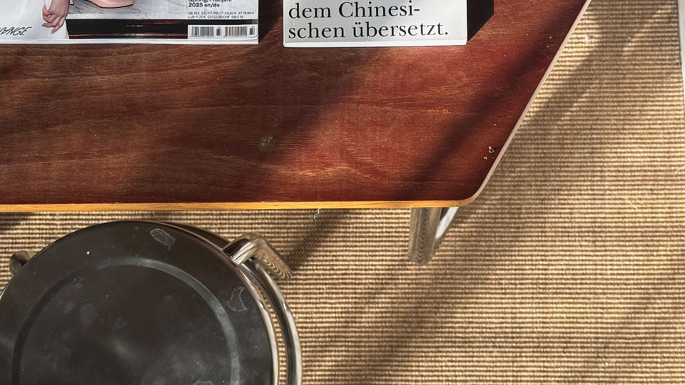 Top-down view of a wooden coffee table on a woven rug, holding a magazine and a small printed quote card. A round black ashtray or tray sits on the rug beside the table, with sunlight casting shadows across the floor.