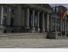 Außenansicht des Reichstagsgebäudes in Berlin mit massiven steinernen Säulen, großen Fenstern und der Inschrift „Dem Deutschen Volke“ über dem Eingang. Im Vordergrund ist ein gepflasterter Platz, mehrere Flaggenmasten und eine Menschenansammlung auf den Treppen sichtbar.