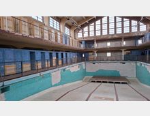 Ehemaliges Hallenbad mit leerem, gefliestem Becken in einem verfallenen Zustand. Die Wände sind teilweise mit blauen und türkisfarbenen Fliesen bedeckt, im oberen Bereich befinden sich alte Umkleidekabinen und eine umlaufende Galerie mit Geländern, darüber große Fensterflächen.