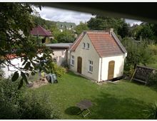 Ein kleiner Garten mit einer gelb gestrichenen Gartenhütte, die ein rotes Ziegeldach und Holztüren hat. Im Vordergrund steht ein runder Holztisch mit einem Stuhl, umgeben von Rasen und Bäumen; im Hintergrund sind weitere Häuser und Grünflächen zu sehen.