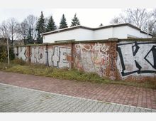 Backsteinmauer mit Graffiti, davor ein Gehweg mit Pflastersteinen und etwas überwuchertes Gebüsch, im Hintergrund ein flaches, weißes Gebäude und kahle Bäume sowie einige Nadelbäume.