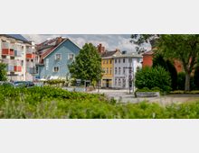 Straßenszene mit mehrstöckigen Gebäuden, darunter ein blaues Gebäude mit „Hotel“-Schriftzug, gelbe und weiße Häuser sowie Balkone mit Blumenkästen. Im Vordergrund befindet sich ein Bereich mit Büschen und Bäumen.