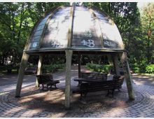 Rundes Pavillon mit einem gewölbten Dach aus Metallplatten, das von vier schrägen Betonpfeilern getragen wird, umgeben von Parkbänken und Bäumen in einem schattigen Waldpark. Das Dach ist mit Graffiti besprüht, und der Boden ist mit Kopfsteinpflaster ausgelegt.