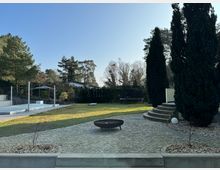 Ein gepflegter Garten mit einer gepflasterten Terrasse, auf der eine runde Feuerschale steht, und einer halbkreisförmigen Treppe, die zu einer erhöhten Fläche führt. Im Hintergrund gibt es einen Rasen, hohe Zypressen, vereinzelt Bäume, ein Trampolin und ein überdachtes Pool mit Schienen.