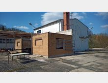 Ein industrielles Gelände mit einem flachen, gelben Backsteingebäude und einem weißen Gebäude im Hintergrund mit Wandzeichnung einer Hand. Der Betonboden ist teilweise verwittert, und mehrere freistehende Tische stehen unter einer Überdachung.