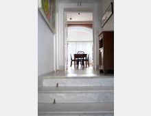 Blick durch einen schlichten Flur mit weißen Wänden und Marmortreppe zu einem hellen Esszimmer mit Holztisch und Stühlen, vor großen Fenstern mit halbtransparenten Vorhängen.