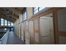 Ein verlassener Gebäudeinnenraum mit verwitterten Umkleidekabinen aus Holz, abblätternder Farbe und hohen Fenstern mit roten Rahmen, die Tageslicht hereinlassen. Die Decke hat freiliegende Träger, und rechts verläuft ein Gang entlang einer offenen Galerie mit Geländer.