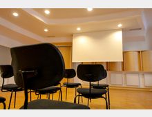 Ein Seminarraum mit schwarzen Stühlen in Reihen, einer weißen Projektionsleinwand an der Wand und einer Deckenbeleuchtung. Der Raum hat eine Holzverkleidung und ein perforiertes Deckenmuster.