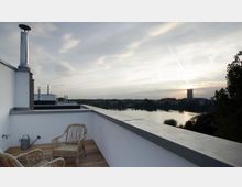 Dachterrasse mit Holzboden, zwei Rattanstühlen und einer Gießkanne, umgeben von weißem Geländer; im Hintergrund ein See, Bäume und eine städtische Skyline bei Sonnenuntergang.