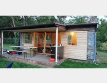 Kleines Gartenhaus mit Holzverkleidung, teilweise renoviert, mit überdachter Terrasse. Davor stehen Bierbänke und ein Tisch, im Hintergrund sind Bäume und eine Wiese zu sehen.