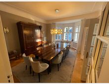 Esszimmer mit einem langen Holztisch, umgeben von hell gepolsterten Stühlen, in einem Raum mit großen, halbkreisförmigen Fenstern und weißen Vorhängen. Im Hintergrund steht ein massiver Holzschrank, und eine moderne Deckenlampe hängt über dem Tisch.