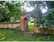 Ziegelsteinbogen mit Holztür in einer historischen Gartenmauer, umgeben von grünen Bäumen und Rasenfläche, mit Blick auf einen sonnigen Garten im Hintergrund.
