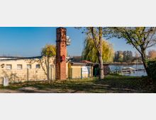 Gelb gestrichenes Gebäude mit Flachdach und einem roten Schornstein, daneben ein Schild mit der Aufschrift „Biergarten“. Im Vordergrund stehen Bäume, und im Hintergrund ist ein See mit Bootsanlegern und Schilfbewuchs zu sehen.