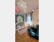 Kinderzimmer mit Holzfußboden, Hausbett und grüner Akzentwand mit großem Waldtier‑Wandbild sowie Wimpelgirlanden und einer weißen Deckenlampe. Ein großes Fenster mit halbtransparenten Vorhängen, ein kleines Regal und ein dunkelgrüner Sessel ergänzen die Einrichtung.