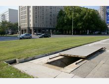 Eine städtische Szene mit einem großen Gebäude im Hintergrund, das eine helle Fassade und zahlreiche rechteckige Fenster hat, sowie einer Straße mit Autos davor. Im Vordergrund befindet sich eine Grünfläche und ein offener, abgesicherter Einstiegsschacht mit einer metallüberdachten Rampe.