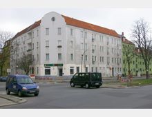 Eckgebäude mit hellgrauer Fassade, roten Ziegeldächern und mehreren rechteckigen Fenstern, im Erdgeschoss befindet sich eine Pizzeria. Im Vordergrund eine Straßenkreuzung mit parkenden Autos und Baustellenabsperrungen.