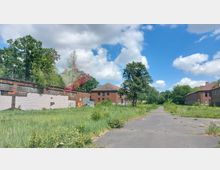 Ehemaliges Industrie- oder Kasernengelände mit verfallenen Backsteingebäuden, teils mit Graffiti bedeckt, umgeben von überwucherndem Grün und vereinzelten Bäumen. Ein asphaltiertes, teilweise beschädigtes Wegstück führt durch die Landschaft, unter einem bewölkten Himmel mit sonnigen Abschnitten.