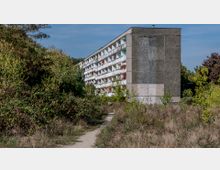 Plattenbau-Wohngebäude mit fünf Etagen und Balkonen, umgeben von dichter Vegetation und einem schmalen Trampelpfad im Vordergrund; Fassade teils grau und teils mit bunten Balkonen gestaltet.