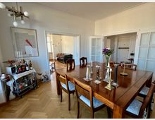 Esszimmer in einer Wohnung mit einem großen hölzernen Esstisch und sieben Stühlen. Auf dem Tisch befinden sich Kerzenhalter und ein Blumenstrauß; im Hintergrund sind ein Barwagen, ein modernes Kunstwerk an der Wand, und ein Durchgang zu einem angrenzenden Wohnzimmer mit einem Ledersessel und Stehlampe zu sehen.