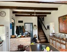 Wohnküche mit rustikalem Charme, geprägt von freiliegenden Holzdeckenbalken, einer Backstein-Trennwand und einem alten Holztresen. Im Hintergrund eine Holztreppe, neben der eine Garderobe, verschiedene Dekorationsgegenstände und ein antiker Spiegel zu sehen sind.