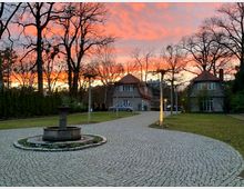 Siedlung mit gepflastertem Weg, einem Brunnen im Vordergrund und zwei Wohnhäusern mit roten Ziegeldächern, umgeben von Bäumen und Rasenflächen; im Hintergrund ein farbenprächtiger Sonnenuntergang.