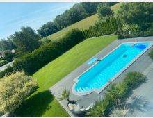 Rechteckiger Außenpool mit grauer Steinumrandung in einem gepflegten Garten mit großem Rasen. Umgeben von Hecken, Sträuchern und kleinen Palmen, mit Blick auf einen bewaldeten Hang und offene Wiesen im Hintergrund.