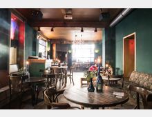 Gemütliches Café oder Bar mit Holztischen und -stühlen sowie einer grünen Wandgestaltung. Im Raum befinden sich Kerzen und Blumen auf den Tischen, eine Theke im Hintergrund, und ein großes Fenster sorgt für Tageslicht.