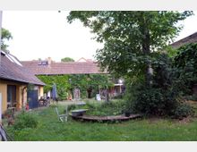 Grüner Innenhof mit großen Bäumen, einer Wiese und Beeten, umgeben von Gebäuden mit gelben und mit Efeu bewachsenen Fassaden sowie Ziegeldächern. Im Hof stehen Gartenmöbel und ein altes Metallbecken auf einem Holzpodest.