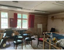 Ein einfacher Raum mit hohen Fenstern und roten Vorhängen, einer Backsteinwand und unvollendeten Möbelprojekten. Auf dem Boden stehen Stühle, während ein Tisch mit Werkzeugen, Farbdosen und anderen Materialien belegt ist.