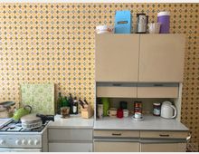 Küche mit Retro-Design aus den 1970er Jahren, gekennzeichnet durch gelb-orange gemusterte Tapete. Zu sehen sind cremefarbene Hängeschränke, ein Gasherd und eine Arbeitsfläche mit Geschirr, Flaschen, Kochutensilien und einem Wasserkocher.