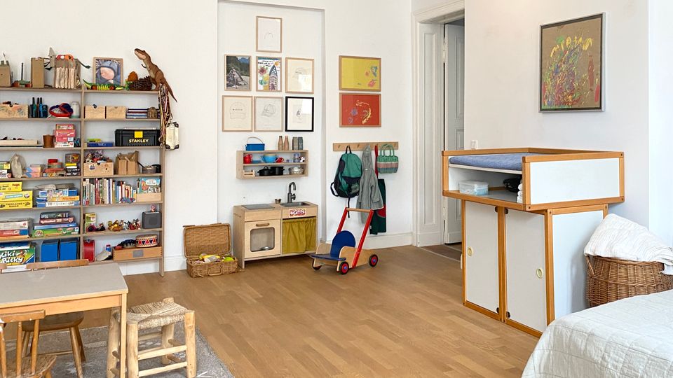 Heller, weiß gestrichener Spiel- und Wohnraum mit Holzfußboden, niedrigem Tisch und kleinen Stühlen sowie offenen Regalen voller Spielzeug und Bücher. An der Wand hängen mehrere gerahmte Bilder über einer Kinder-Spielküche; daneben Garderobenhaken und ein offener Durchgang zu einem Flur, rechts ein Wickel- oder Aufbewahrungsmöbel.