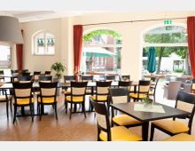 Heller Gastraum mit Holzboden, schwarzen Tischen und Stühlen mit gelber Polsterung und gemusterten Rückenlehnen, große Fenster mit Rundbögen und Blick auf einen Außenbereich mit Sonnenschirmen und Bäumen.