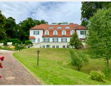 Große zweistöckige Villa mit weißer Fassade, roten Ziegeldach und blauen Fensterläden, umgeben von einer grünen Rasenfläche und Bäumen, mit einem gepflasterten Weg im Vordergrund.