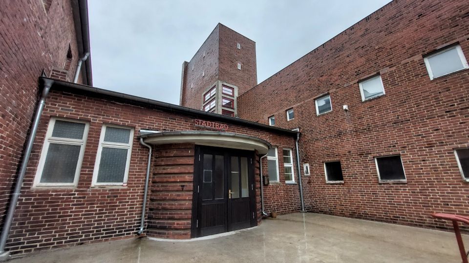 Backsteingebäude eines Stadtbades mit einem Eingang in der Mitte, gekennzeichnet durch eine schwarze Tür und ein rundes Vordach. Umgeben von schlichten Fenstern und hohen, roten Backsteinwänden.
