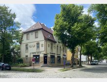 Zweistöckiges Gebäude im traditionellen Stil mit hellem Putz und einem roten, schrägen Dach, das ein Fotogeschäft und eine Drogerie im Erdgeschoss beherbergt. Das Gebäude steht an einer gepflasterten Straße, umgeben von großen, grünen Bäumen bei sonnigem Wetter.