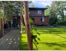 Holzhaus mit zwei Etagen in einem Garten mit grünem Rasen und Pflastersteinen, links ein mit Holzgestellen überdachter Weg. Im Hintergrund stehen Gartenmöbel und Bäume.
