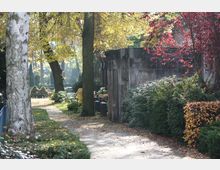 Ein parkähnlicher Friedhof mit herbstlicher Atmosphäre, schattigen Bäumen und buntem Laub. Rechts im Bild ist ein historisches Sandsteinmausoleum von Büschen und rot-goldenen Blättern umgeben.
