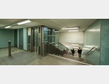 Ein unterirdischer U-Bahn-Zugang mit Treppen, einem gläsernen Aufzug auf der linken Seite und türkisfarbenen Wandfliesen. Zwei Personen gehen die Treppe hinunter in Richtung eines Bahnsteigs mit Hinweisschildern.