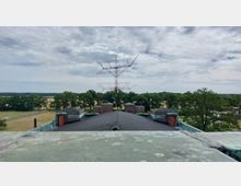 Blick auf eine historische Funkanlage mit einem großen roten Sendemast im Zentrum, umgeben von mehreren Backsteingebäuden und Bäumen in einer ländlichen Umgebung unter wolkigem Himmel. Im Vordergrund ist ein Teil eines grünlichen Metalldachs sichtbar.