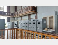 Innenraum eines historischen Maschinengebäudes mit großen grauen Schalttafeln und technischen Geräten. Die hohen Fenster, eine Holzbalkonbrüstung und Backsteinelemente prägen die Architektur.