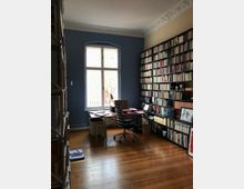 Ein Arbeitszimmer mit Holzboden, einem großen Fenster mit blau gestrichener Wand, einem Schreibtisch mit Bürostuhl und Bücherregalen, die die rechte Wand vollständig bedecken. Die Decke ist mit dekorativen Stuckdetails verziert.