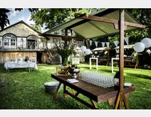 Grüne Außenfläche mit dekorierten Festständen, Picknicktischen und weißen Lampions; im Hintergrund ein zweistöckiges Holzgebäude mit Balkon und Treppe. Im Vordergrund ein Getränketisch mit Gläsern und gekühlten Flaschen.