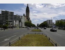 Breite Straße in einer städtischen Umgebung mit zentraler Grünfläche, flankiert von mehrspurigen Fahrbahnen. Im Hintergrund ist ein hoher Kirchturm sichtbar, umgeben von modernen und historischen Gebäuden sowie einer U-Bahn-Station auf der linken Seite.