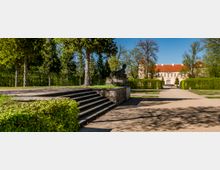Formal garden with a wide gravel path bordered by trimmed hedges and tall trees, featuring stone steps and a statue on a raised terrace. In the background, a large historic palace-like building with a red tiled roof stands under a clear blue sky.