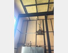 Innenansicht eines Raums mit gelb-beigen Wänden und schwarzem Holzfachwerk. Im Vordergrund ist ein silberner Metalltank mit angeschlossenen Rohren und Ventilen zu sehen, darüber verlaufen Kabel und eine kleine Box an der Wand.