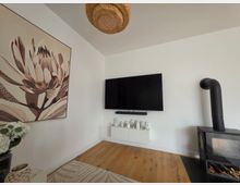 Helle Wohnzimmerecke mit weiß gestrichenen Wänden, Holzboden und einem an der Wand montierten Flachbildfernseher über einem schwebenden Lowboard mit kleinen Deko-Häusern. Links hängt ein großes Blumenbild, rechts steht ein schwarzer Kaminofen mit Ofenrohr; an der Decke hängt eine geflochtene Rattanlampe.